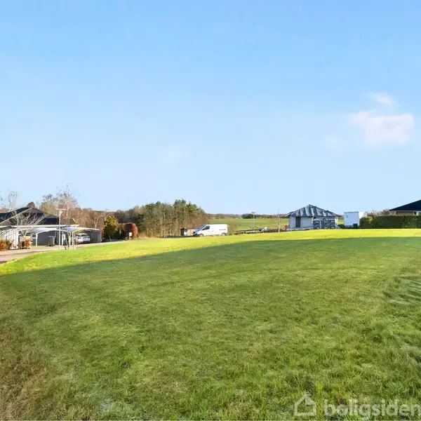 Græsplæne ligger stille i et åbent boligområde med flere huse og en vej i baggrunden under en lys blå himmel.