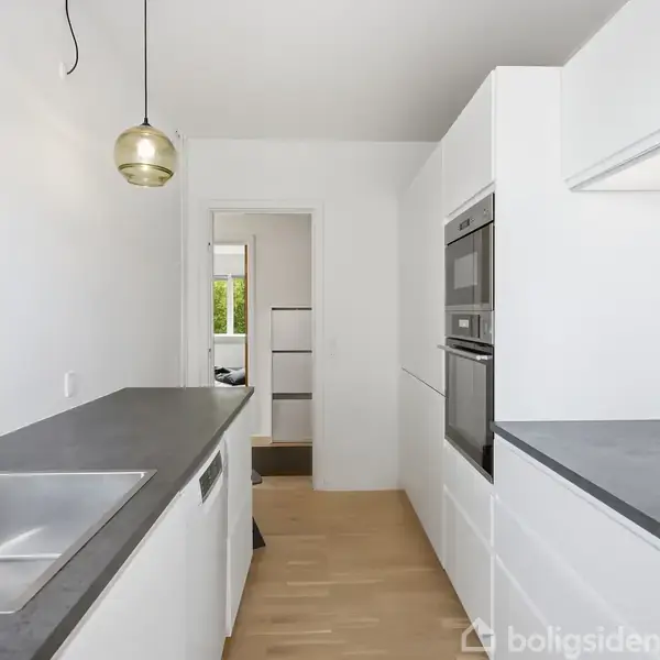 Et moderne køkken har hvide skabe, stålvaske og indbyggede ovne. En lampe hænger over bordpladen. I baggrunden ses en døråbning med udsigt til grønne omgivelser.