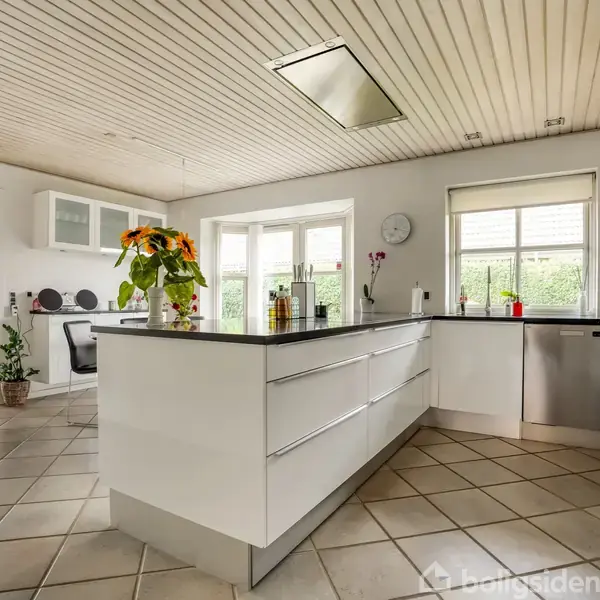 Et moderne køkken med en stor, hvid køkkenø, omgivet af skabsplads og integrerede apparater. Sollys strømmer ind gennem vinduerne, og blomster pynter bordet. En spisestue kan ses i baggrunden.