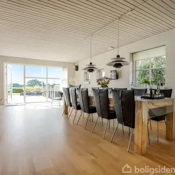 Spisebord med sorte stole står i spisestue. Ved siden af er glasdøre, der åbner ud til terrasse med udsigt over haven og blå himmel. Lampen hænger over bordet.