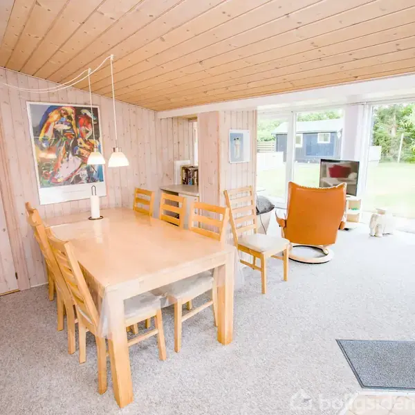 Træspisebord med seks stole står i et lyst rum. Ved siden af er en orange lænestol med udsigt til en have gennem store vinduer. Maleri hænger på væggen.