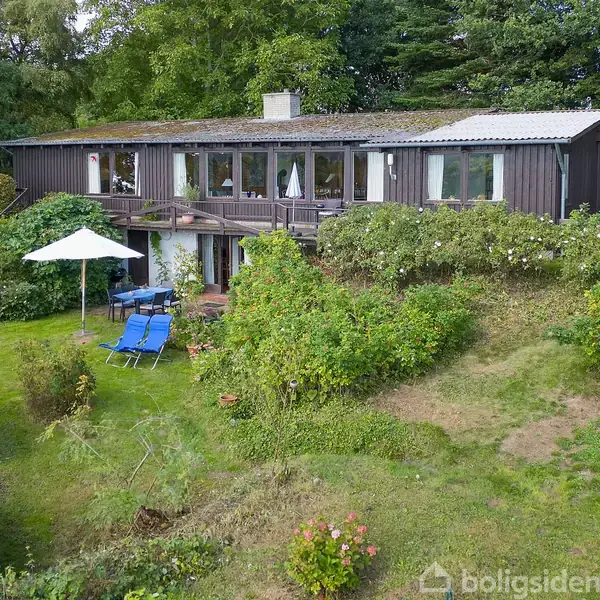 Et mørkt træhus med store vinduer står midt i en frodig have. Foran huset er der hvide havemøbler og en hvid parasol. Grønt løv omgiver området.