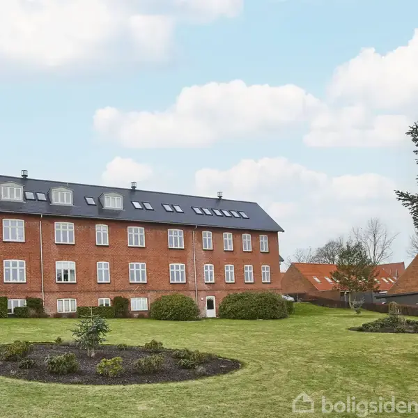 En tre-etagers murstensbygning med sorte tegltag er omgivet af en veldrevet grøn have med blomsterbede. Andre bygninger og træer ses i baggrunden under en blå himmel med skyer.