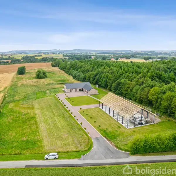 Hus omgivet af græsmarker og skovområder, ligger på en lang vej med en parkeret bil. Store åbne marker og en klar blå himmel danner baggrundsscenen.