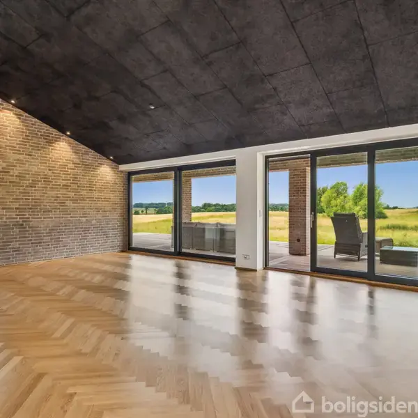 Tomt rum med trægulv og murstensvægge, set indefra. Store vinduer og en glasdør viser en terrasse med havemøbler og en grøn mark udenfor under en blå himmel.