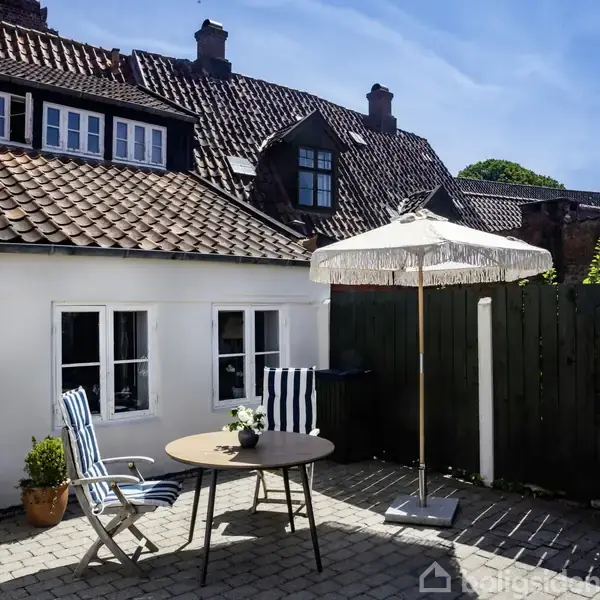 Et rundt bord med to blåstribede stole står under en parasol i en brostensbelagt gårdhave. Baghuset har rødt tegltag og hvide vinduer mod en blå himmel.