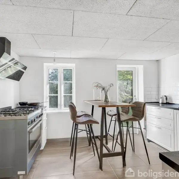 Et moderne køkken med en gaskomfur til venstre og en indbygget emhætte over. En høj bord og tre stole står midt i rummet. Store vinduer giver naturligt lys.