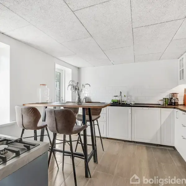 Et moderne køkken med hvide skabe står i et lyst rum. Der er en barstol ved siden af et højt bord. Store vinduer lader naturligt lys skinne ind.