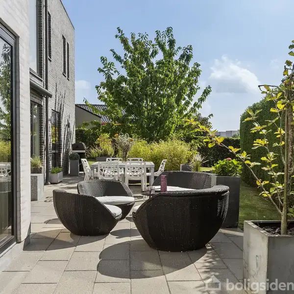 Runde fletstole står på en flisebelagt terrasse ved et moderne hus. En stor træstolgruppe ses længere væk, omgivet af tætte buske og et blomstrende træ under en blå himmel.