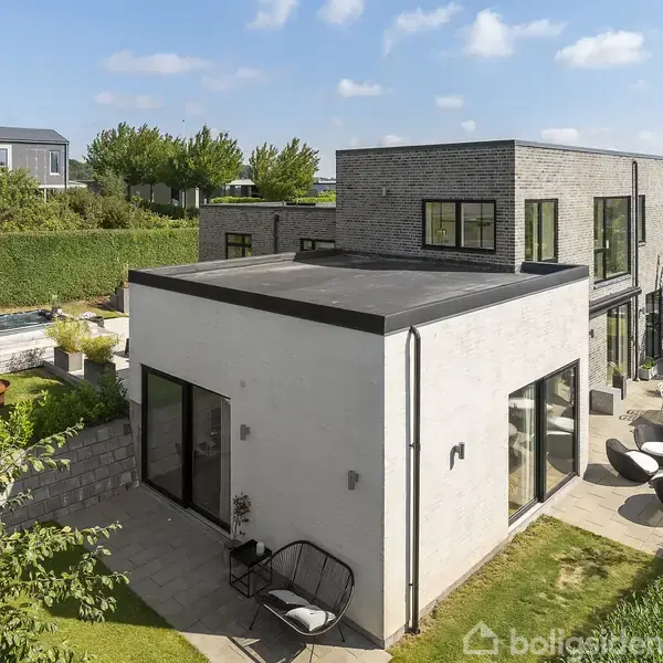 En moderne, kubisk villa i to etager med store vinduer og fladt tag, står i en velplejet have med terrasse og havemøbler, omgivet af grønt og hække.