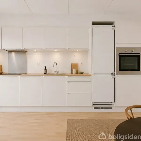 Hvidt køkken med indbyggede skabe og apparater. Ovnen er placeret ved siden af et stort køleskab. På bordet står grønne planter. Lysindfald skaber en lys og rummelig atmosfære.