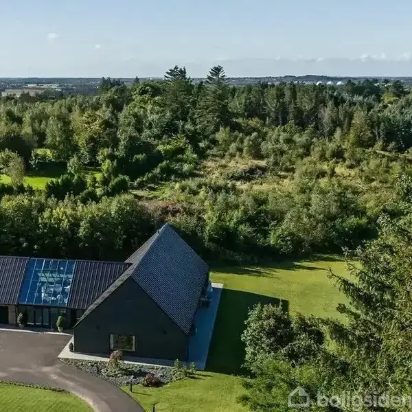 Et moderne hus med sort tag står i centrum, omkranset af en velplejet græsplæne og tæt skovområde, under en klar blå himmel.