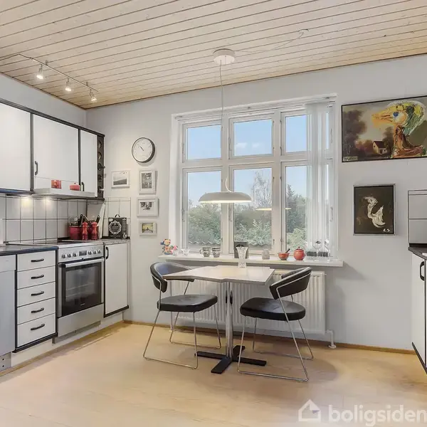 Køkken med hvide skabe og sorte håndtag, bord med to stole under en lampe ved vindue. Vægge dekoreret med billeder. Køkkenet har komfur, opvaskemaskine og et ur.