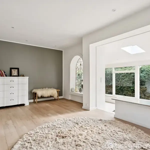 Et lyst rum med en hvid kommode og bøger mod en grå væg. Et rundt tæppe ligger på trægulvet, ved siden af et stort vindue med udsigt til en have.