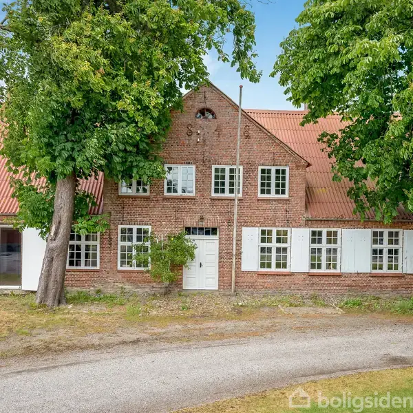 En stor, rød murstensbygning med rødt tegltag og hvide vinduer står mellem to store træer; en grusvej løber foran bygningen, og grønt græs omkranser området.