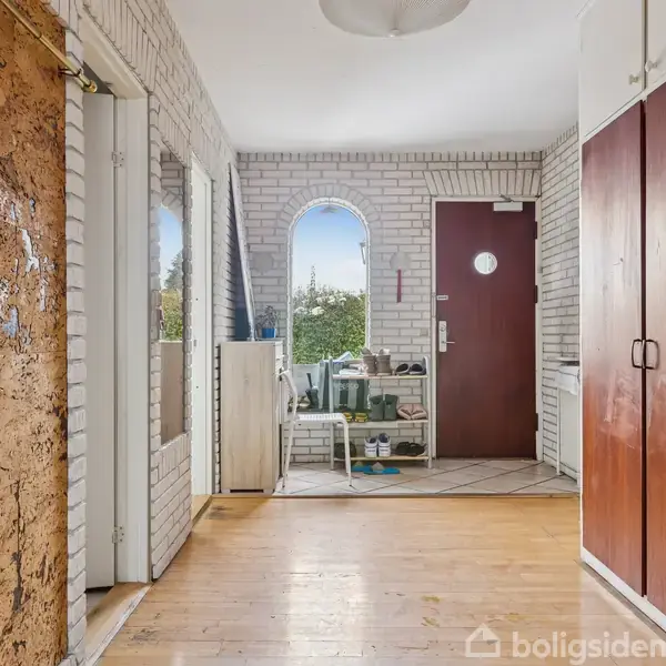 En entre med hvide murstensvægge og et trægulv. En brun dør med rundt vindue sidder til højre, mens venstre side har spejlet skabslåger. Møbler er sparsomt placeret.