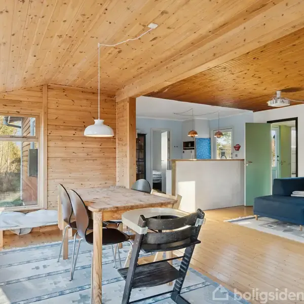 Et åbent køkken-alrum med trævægge og loft, indeholder et spisebord med stole og en sofa ved siden af. Store vinduer lader sollyset strømme ind, hvilket skaber en lys atmosfære.