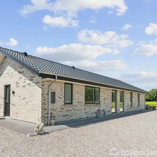 Et hvidt murstenshus står stille på en grusbelagt gårdsplads omgivet af grønt landskab og spredte træer under en blå himmel med få skyer.
