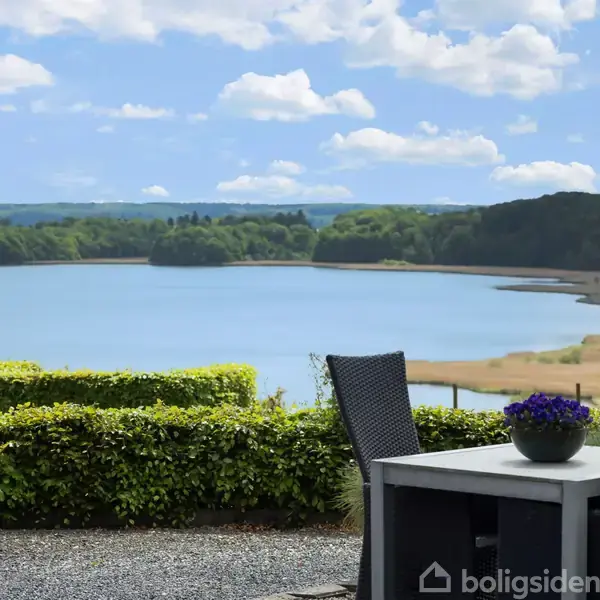 Et par sorte stole står omkring et gråt bord med en blå blomsterpotte på. Udsigt over en sø omgivet af træer, under en blå himmel med skyer.
