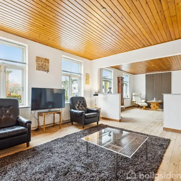 Stue med sort læderstol og sofa, stående foran fjernsyn og glasbord på gråt tæppe. Trægulv og træloft, lyse vægge og store vinduer, der slipper naturligt lys ind.