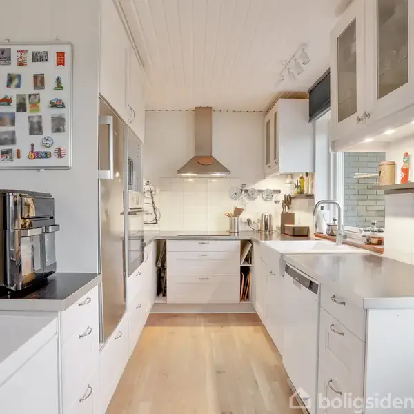 En moderne køkken indrettet med hvide skabe og skuffer. Der ses en espressomaskine på bordet, og køkkenet har indbygget komfur og emhætte. Hylderne indeholder diverse køkkenartikler og madvarer.