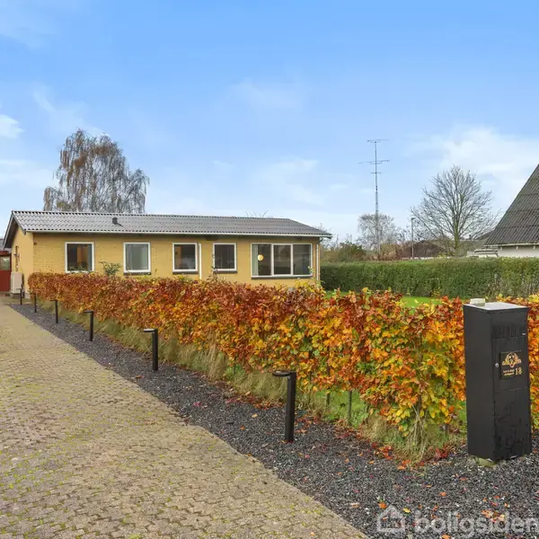 En gul murstensvilla med fladt tag står bag en hæk med efterårsfarver i et parcelhuskvarter. En til salg skilt ved hækken har teksten "Mæglerhuset til salg".