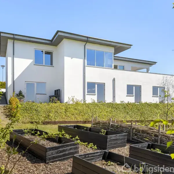 Hvid, moderne to-etagers hus står i en have med hævede blomsterbede og grønne buske. Klar blå himmel skaber en solrig og indbydende atmosfære.