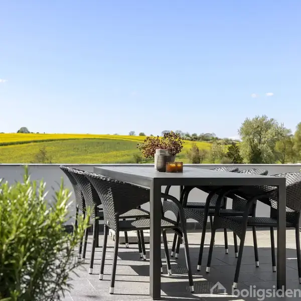 En sort spisebord med sorte stole står på en terrasse. Der er planter i krukker omkring bordet, og baggrunden viser et landskab med gule marker og træer under en blå himmel.