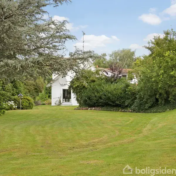 Hvidt hus står stille omgivet af frodig have og grønne træer under en blå himmel.