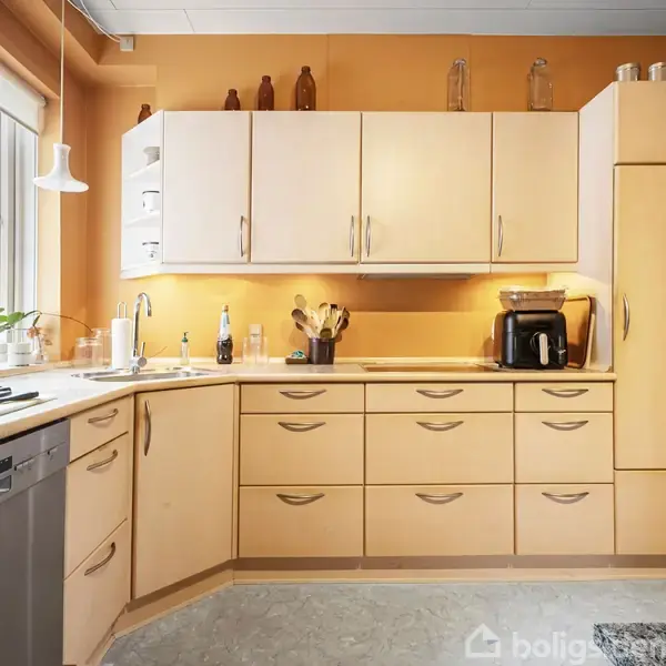 Køkken med lyse træskabe, udfører opbevaring. Køkkenmaskiner og køkkenredskaber står på bordpladen. Et stort vindue til venstre giver naturligt lys, og gulvet har et lyst mønster.