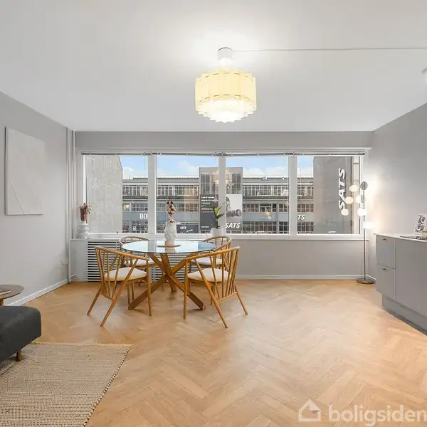 Spisebord står i centrum med fire stole omkring det, placeret i en lys stue med trægulv og store vinduer. En sofa og skænk udgør stuens møblering.