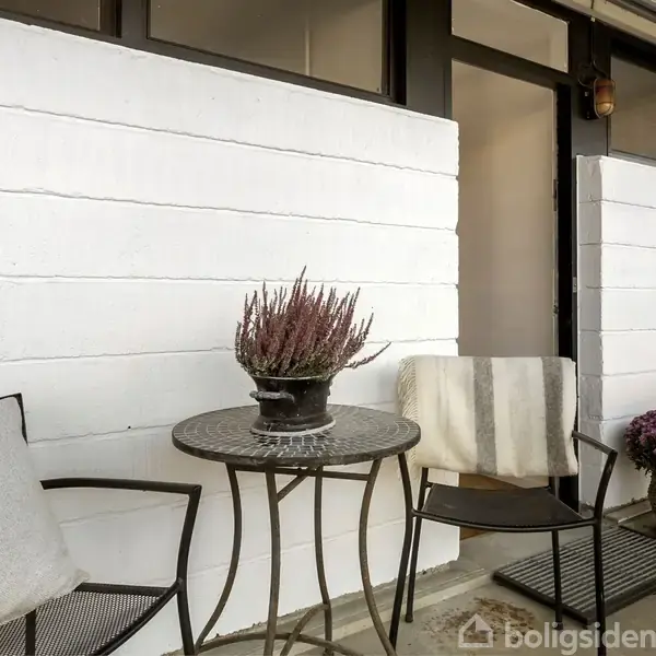 Et rundt bord med en plante ovenpå står mellem to stole ved en hvid murstensvæg, i en udendørs terrasseindretning med tæppe og ekstra plantearrangementer.