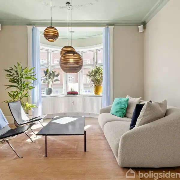 Sofa og lænestole omkranser et lavt sofabord i en stue med trægulv. Lysekrone hænger fra lysegrøn loft. Store vinduer med planter udsigt til murstensbygninger udenfor. Ingen tekst.