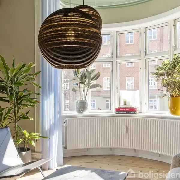 Pendellampe hænger over en vindueskarm med planter i farverige krukker. I baggrunden ses en murstensbygning. Tre bøger er stablet: "The Magic of Thinking Big," "Four Thousand Weeks," "Think Again."