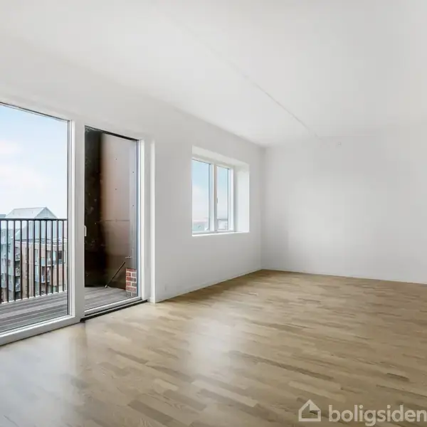 Et tomt rum med trægulv og hvidmalede vægge åbner op mod en balkon gennem en glasdør. Store vinduer giver udsigt til en klar himmel og omkringliggende bygninger.