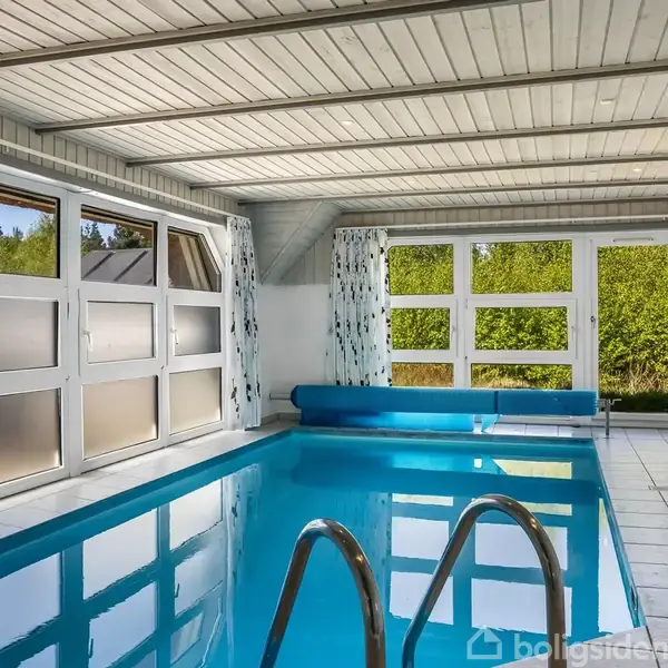 Indendørs swimmingpool står stille i et lyst rum med store vinduer, der giver udsigt til grønne træer udenfor. Hvide fliser omgiver poolen, og lysegardiner hænger ved vinduerne.