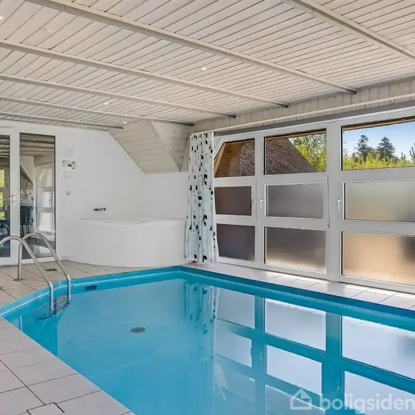 Indendørs swimmingpool med metalkant og trin, fyldt med vand. Store vinduer med udsigt til grøn natur. Fliser dækker gulvet, og der er lyse træplanker på loftet.