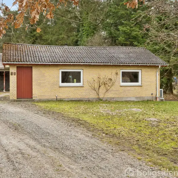 En gul murstensbygning med rød dør og to vinduer står i en have med grusvej. Nummeret "85" er synligt ved hoveddøren. Omkring er der træer og en garage.