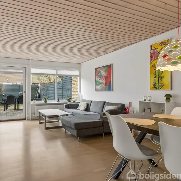 Stue med grå hjørnesofa står mod en hvid væg med farverige malerier. Rundt spisebord med stole under farverig lampe. Store vinduer åbner til terrasse med havemøbler udenfor.