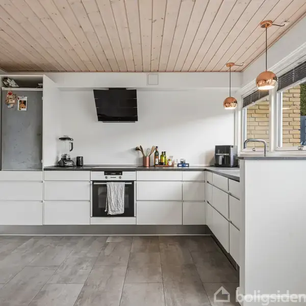 Køkken med hvide skabe og en sort ovn. Kedel og flasker på bordet. Træloft og store vinduer til venstre giver lys. To hængelamper og dør til højre.