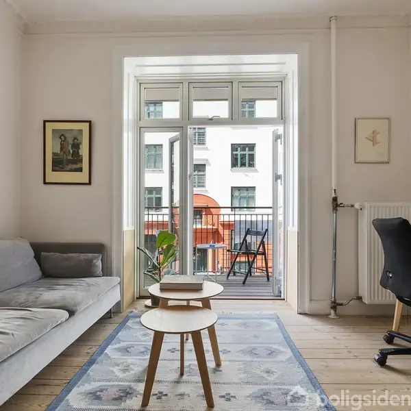 Grå sofa står ved siden af runde borde i et lyst stueområde. Et skrivebord med en computerskærm og stol er placeret i hjørnet nær glasdøre, der fører ud til en altan.