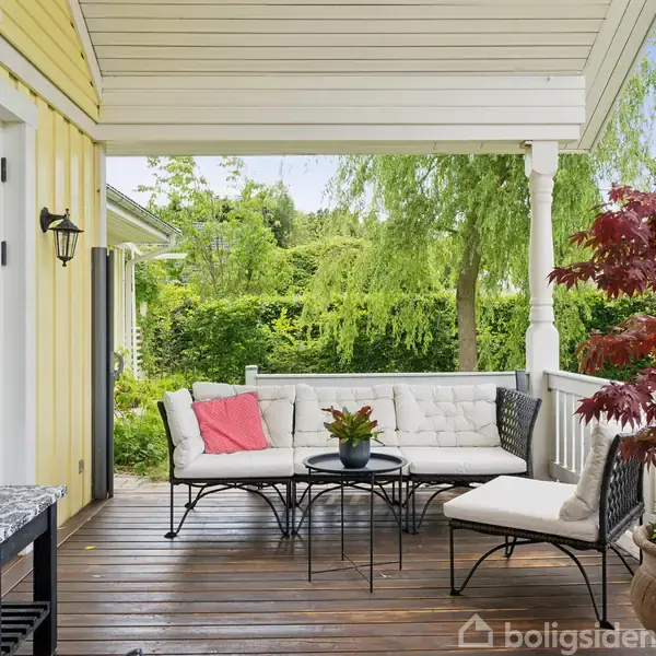 En terrasse med en hvid sofa og et bord med en plante. En stol står ved siden af, og en rød plante i en potte er i forgrunden. Grønt haveområde omringer terrassen.