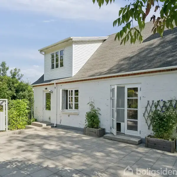 En hvidmalet to-etagers bygning med skråt tag står stille på en flisebelagt terrasse, omgivet af grønne buske og træer under en klar blå himmel.