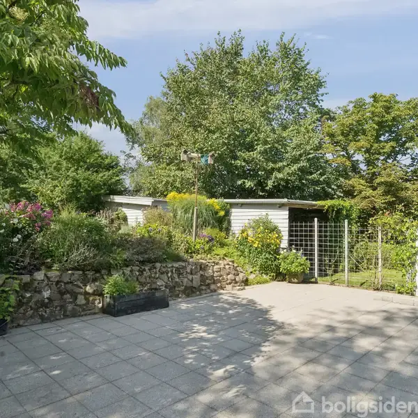 En flisebelagt terrasse står stille i en frodig have med forskellige buske og blomster ved en lav stenmur, tæt på et hvidt skur omgivet af grønne træer og buske.