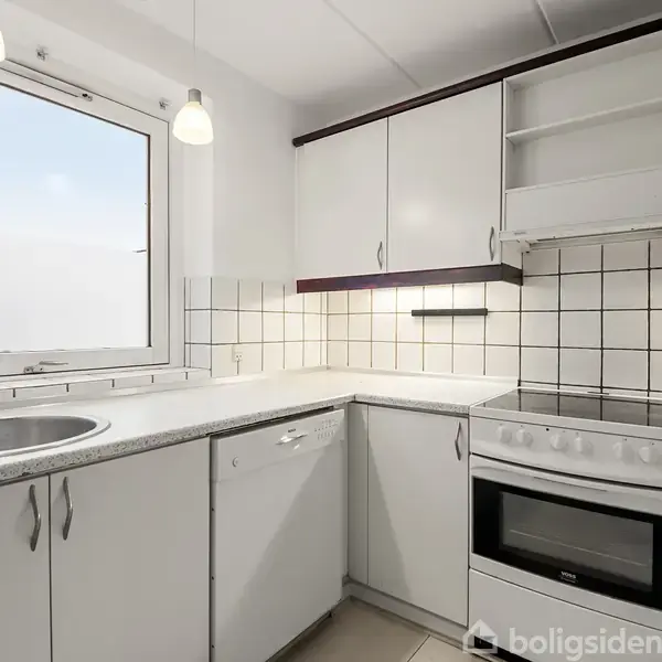 Et moderne køkken med hvide skabe og sorte detaljer. En indbygget ovn og kogeplade ved siden af en vask med rustfri stål. Flisebelagt væg og stort vindue til venstre.