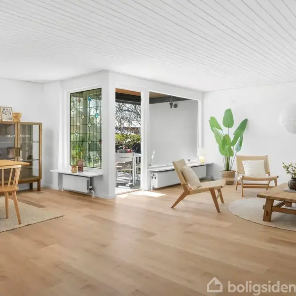 Spisestuemøbler står i et lyst rum med trægulv og hvide vægge. Et rundt bord med planter, stole og en sofa er nær et åbent vinduesparti med udsigt til en have.