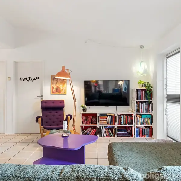 En stue med en lilla stol og et lilla bord foran en grøn sofa. En fladskærms-tv er placeret over en bogreol med farverige bøger. På døren står der "JONATHAN".