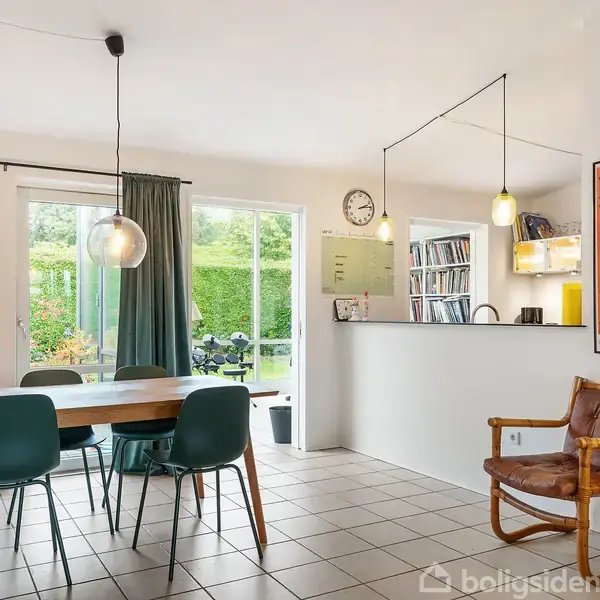 Spisestue med et træbord og seks grønne stole. Lamper hænger over bordet. Til venstre er store vinduer med gardiner, til højre en lænestol, og en "Vertigo"-plakat hænger på væggen.