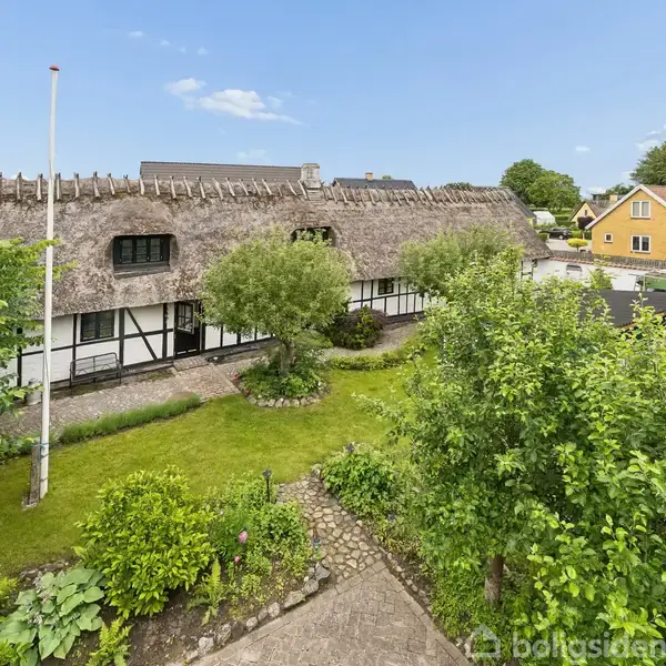 Stråtækt bindingsværkshus omgivet af en velplejet have med stier og træer i en stille nabolag. En flagstang står til venstre, og gule huse ses i baggrunden.
