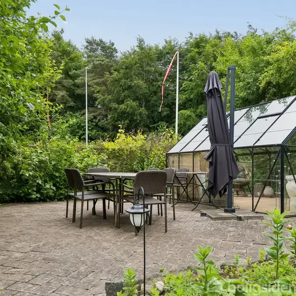 Bindingsværkshus med stråtag står i en grøn have med træer og buske. Haven er omgivet af en stensti, og i baggrunden ses flere huse og en blå himmel.